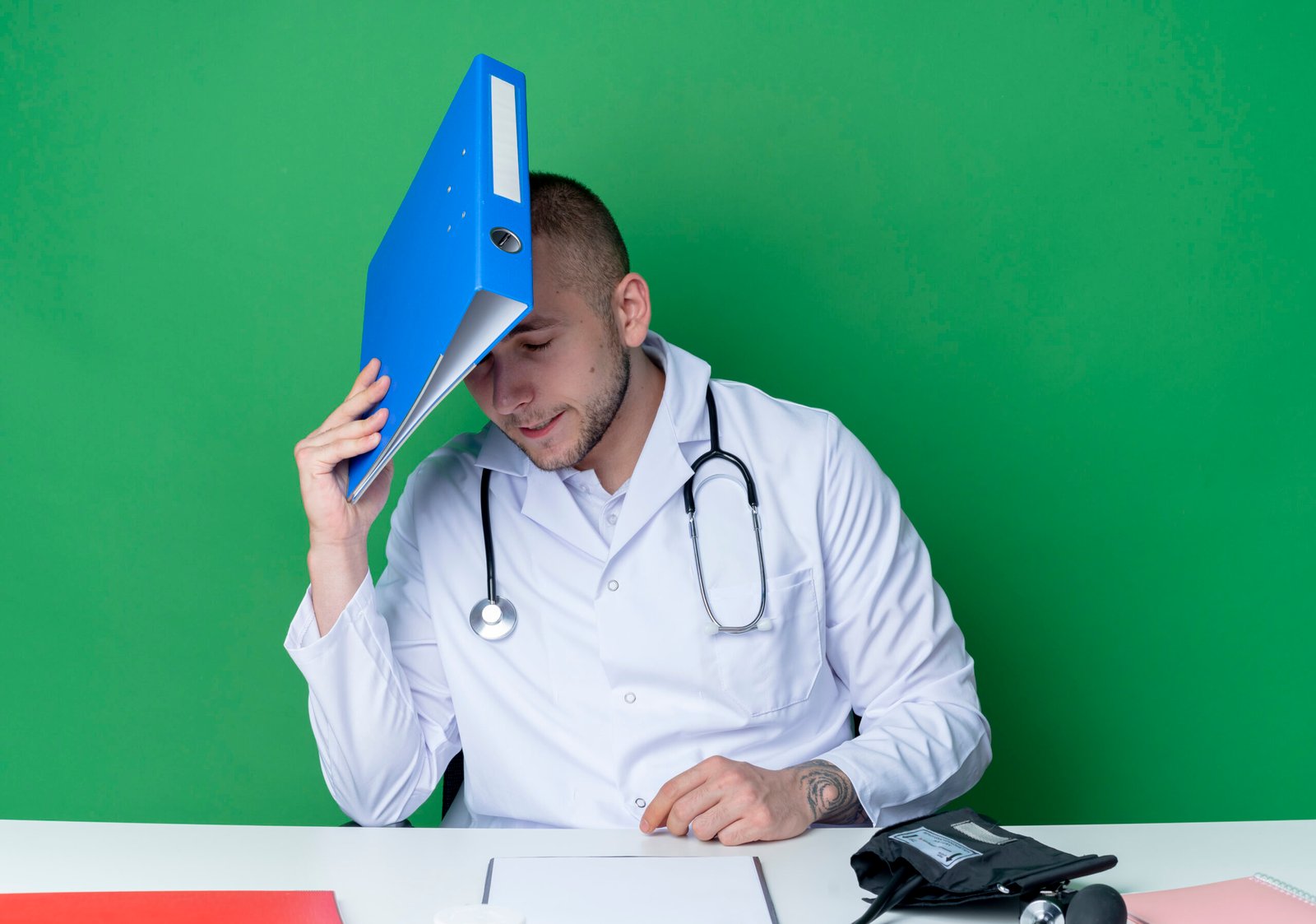 tired young male doctor wearing medical robe and stethoscope sitting at desk with work tools holding folder and touching head with it with closed eyes isolated on green background