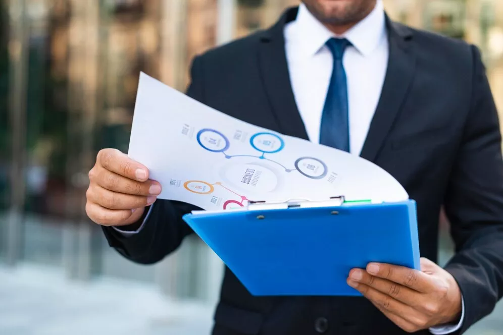A businessman in a suit reviewing a document with a credentialing process flowchart on a blue clipboard outdoors.
